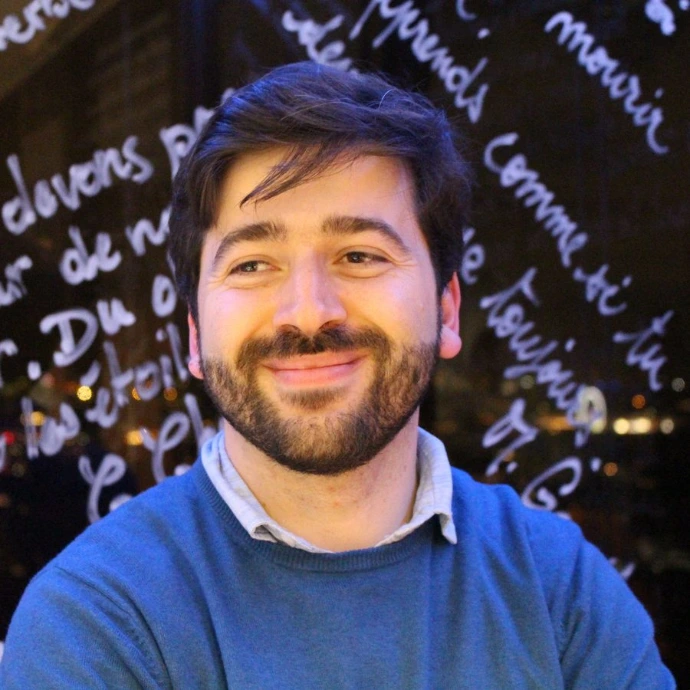 Imagem de um homem branco, de sueter, com um fundo escuro. O Homem de aproximadamente 35 anos, tem cabelo e barba castanhos